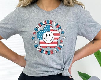 May include: Grey t-shirt featuring a red, white and blue flower design. The flower has a smiley face in the centre and the words "American Vibes" are written around it.