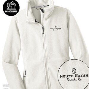 Könnte beinhalten: Weißes, besticktes Fleece-Jacket mit durchgehendem Reißverschluss und Stehkragen. Auf der Jacke ist der Text "Neuro Nurse Sarah, RN" auf der Brust gestickt. Das Bild enthält auch eine runde Grafik mit dem Text "Embroidered" und der Farbe "Winter White".