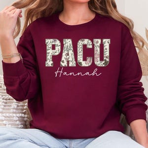 May include: Burgundy sweatshirt featuring the word "PACU" in floral-filled letters and the name "Hannah" in a cursive font. The sweatshirt is a solid burgundy colour.