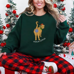 Op de afbeelding: Een donkergroene sweatshirt met een cartoonkat met een kerstmuts en een sneeuwvlok. De tekst "MEOWY CATMAS" staat onder de kat. De persoon draagt een rood-zwarte geruite pyjamabroek.