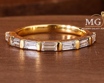 May include: A gold band ring with rectangular, clear gemstones set around the band. The ring is on a wooden surface. The ring has a Modern Galaxy Jewel logo.