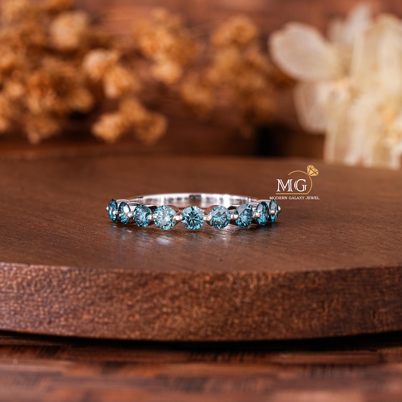 May include: A silver ring with a row of round, light blue gemstones. The ring is set on a dark brown wooden surface, with blurred floral elements in the background. The ring is a piece of jewelry.
