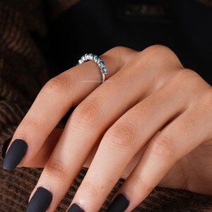 May include: A silver ring with a row of small, round, blue gemstones. The ring is on a person's finger, with dark nail polish. The background is a brown, textured fabric.