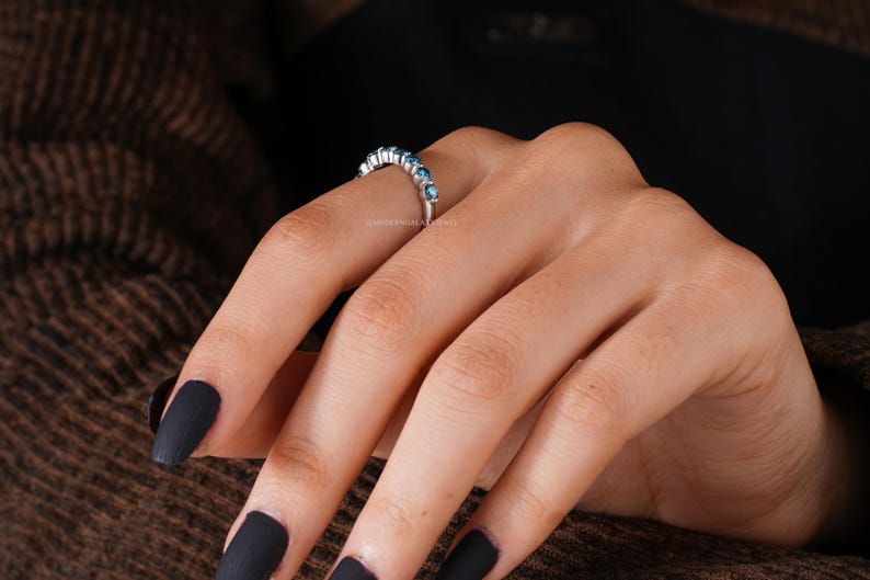 May include: A silver ring with a row of small, round, blue gemstones. The ring is on a person's finger, with dark nail polish. The background is a brown, textured fabric.