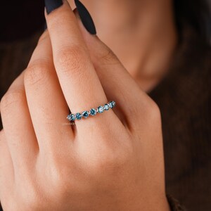 May include: A silver ring with a row of blue gemstones. The ring is worn on a finger, showcasing the gemstones' color and sparkle. The ring's design is simple, with the gemstones set closely together.