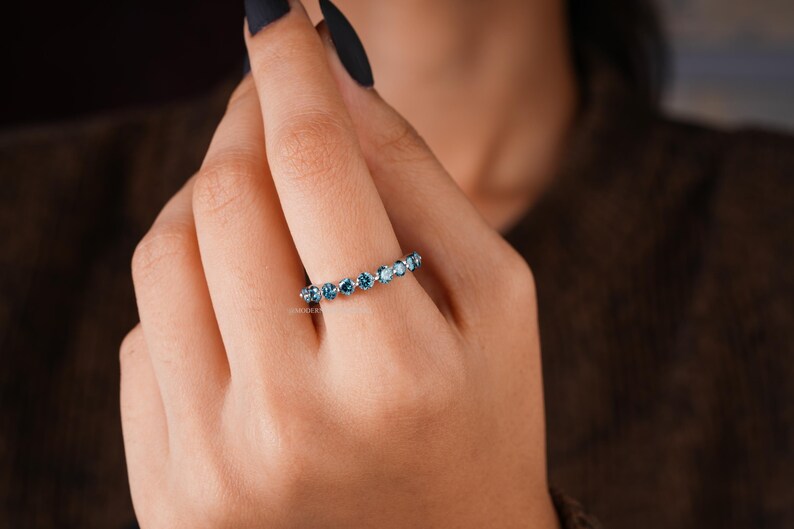 May include: A silver ring with a row of blue gemstones. The ring is worn on a finger, showcasing the gemstones' color and sparkle. The ring's design is simple, with the gemstones set closely together.