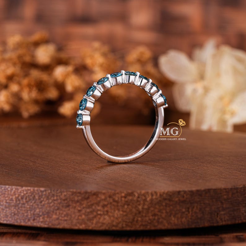 May include: A silver ring with a row of blue gemstones. The ring is set on a wooden surface, with a blurred background of dried flowers and a white flower. The ring has a modern design.