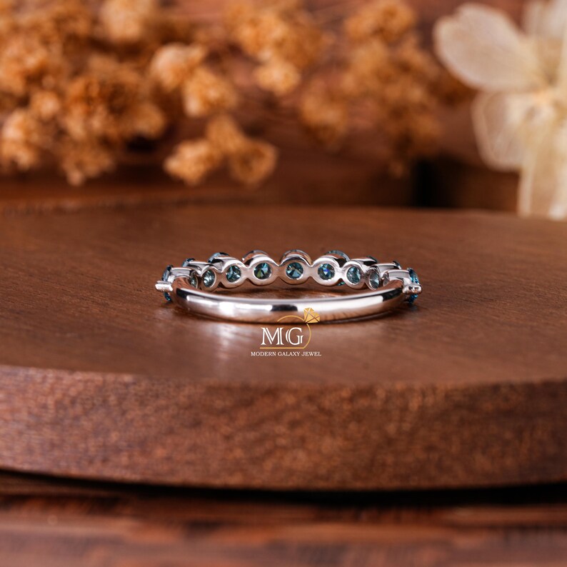 May include: A silver ring with a row of small, round, blue-green gemstones. The ring is set on a wooden surface, with the logo "MG MODERN GALAXY JEWEL" visible. The ring's design is simple and elegant, suitable for everyday wear or special occasions.