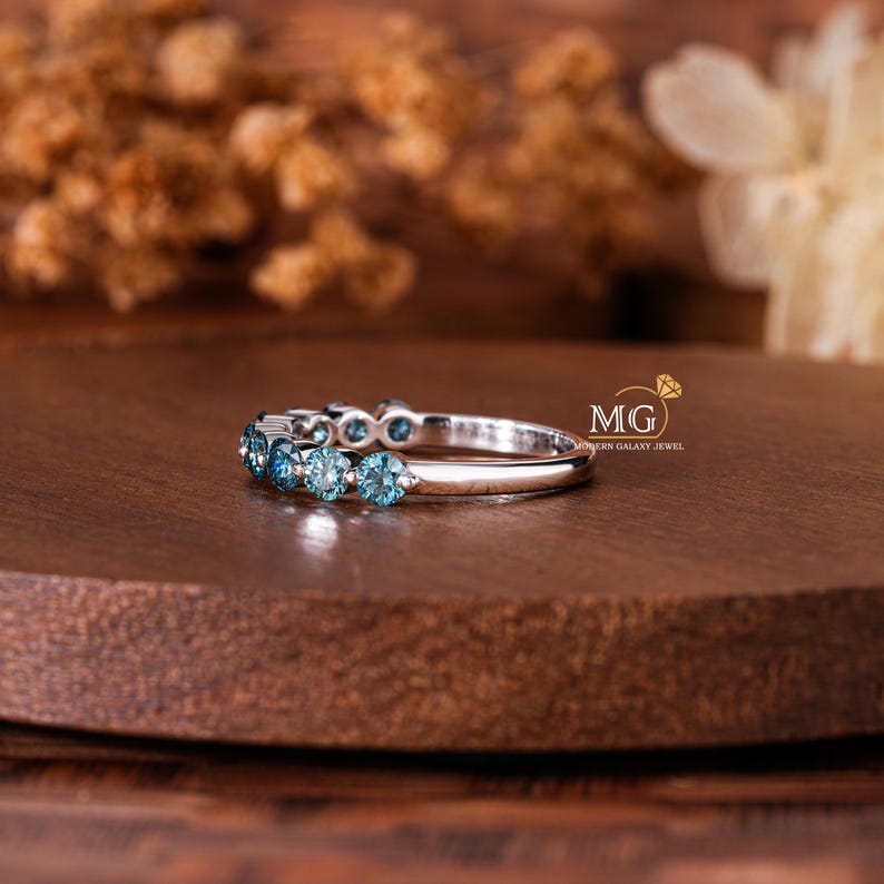 May include: A silver ring with multiple round, light blue gemstones. The ring is set on a dark brown wooden surface, with a blurred background of dried flowers and a white flower.