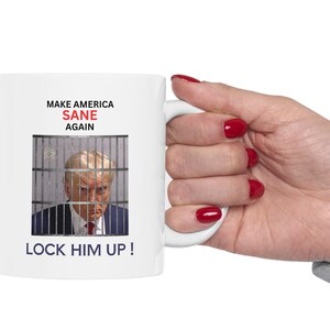 May include: White ceramic mug with the text "MAKE AMERICA SANE AGAIN" and "LOCK HIM UP!". The mug features an image of a person behind bars. The mug is held by a hand with red nail polish.