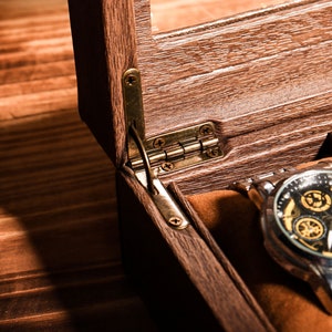 May include: A wooden watch box with a hinged lid, open to reveal a watch with a black face and gold accents. The watch is on a silver metal band.