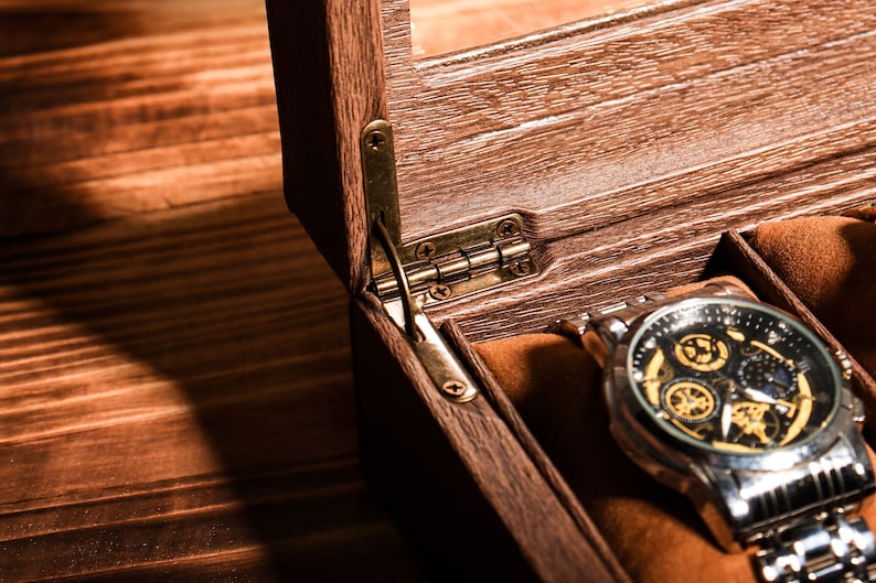 May include: A wooden watch box with a hinged lid, open to reveal a watch with a black face and gold accents. The watch is on a silver metal band.