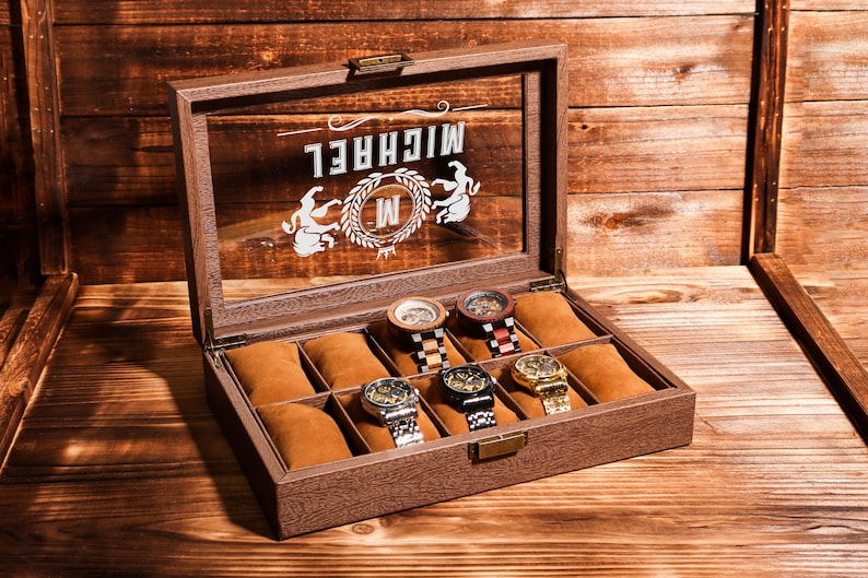May include: A brown wooden watch box with a glass top. The box has a label that says "MICHAEL" with a stylised "M" inside a wreath. The box has six compartments for watches, and five of the compartments have watches in them. The watches are various styles and colours.