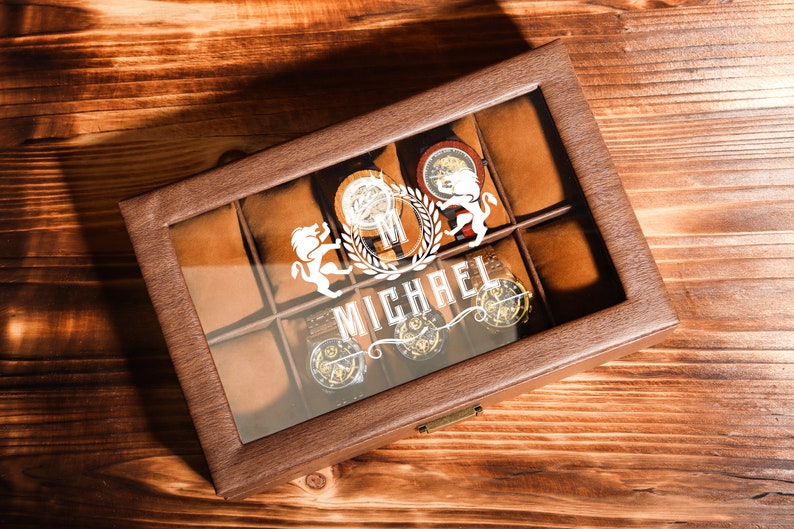 May include: A brown leather watch box with a glass top. The box has eight compartments for storing watches. The box is personalised with the name "MICHAEL" and a lion crest.