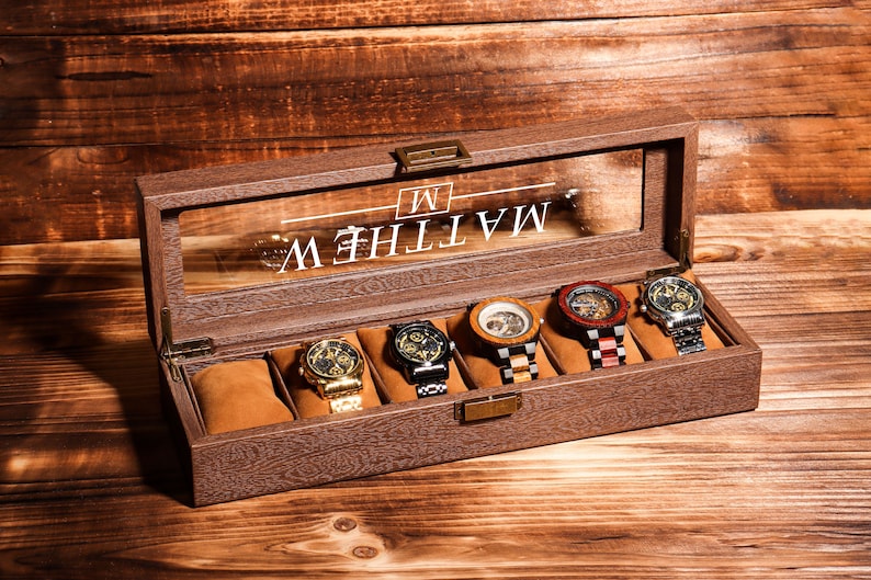 May include: A wooden watch box with a glass top and a brown finish. The box has six slots for watches and is engraved with the name "MATTHEW" and the initial "M".