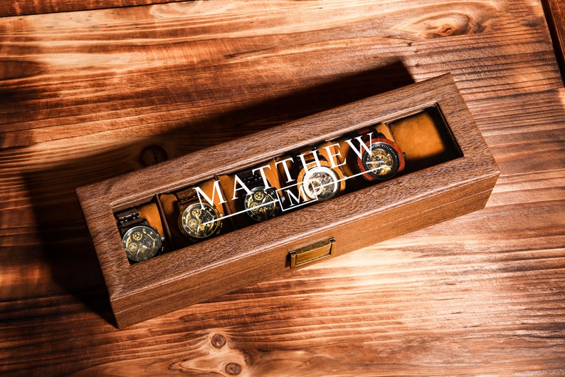 May include: A wooden watch box with a glass top. The box has a personalised inscription that reads "MATTHEW". The box is filled with five watches.
