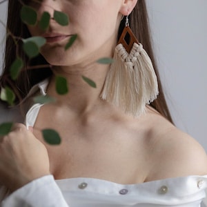 May include: A woman wearing a white button-down shirt with the collar undone, and a pair of brown and white tassel earrings. The earrings have a wooden diamond shape with a hole in the center.