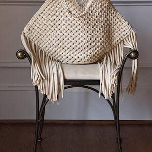 Może przedstawiać: Kremowa torebka z makramy z frędzlami, spoczywająca na krześle z kutego żelaza. Torba ma tkany wzór i uchwyt. Krzesło ma zakrzywioną konstrukcję i neutralne kolorystycznie siedzisko na neutralnym tle.