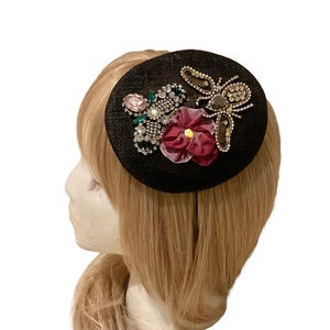 May include: A black fascinator with a decorative design. The design includes a jeweled bee, a cluster of pink and green gems, and a burgundy flower. The fascinator is worn on a mannequin head with blonde hair.