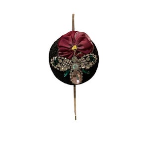 May include: A black hair accessory with a decorative bee design. The bee is embellished with clear, green, and pink gemstones. A burgundy fabric flower with a yellow center sits above the bee. A thin, gold-colored hair stick is attached.