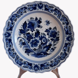 May include: A white ceramic plate with a scalloped edge, adorned with a blue floral design. The plate features a central bouquet of flowers and leaves, with a delicate butterfly accent. The rim is decorated with intricate blue patterns.