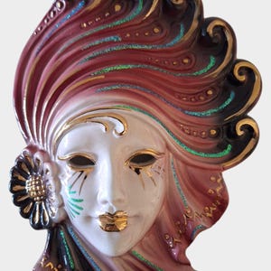 May include: A ceramic wall mask with a white face, gold accents, and a red and black feathered headdress.