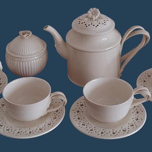 May include: A cream-colored ceramic tea set featuring a teapot with a floral lid, a lidded sugar bowl, and four cups with matching saucers. The saucers and teapot lid have intricate cut-out star patterns, and the cups have curved handles.
