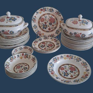 Può includere: Un set di stoviglie in ceramica bianca con un motivo floreale nei colori rosso, blu e giallo. La collezione comprende due zuppiere con coperchio e manici, diversi piatti piani e numerose ciotole, tutti decorati con un motivo floreale centrale.
