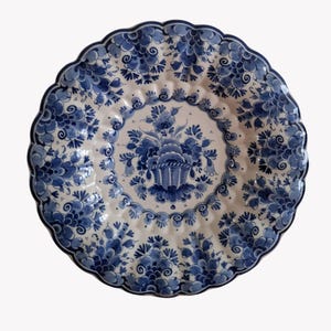 Op de afbeelding: Een ronde, witte keramische plaat met een geschulpte rand, versierd met een blauw bloemmotief. Het midden toont een mand met bloemen, omringd door blauwe patronen. De rand van de plaat is ook versierd met blauwe bloemmotieven.