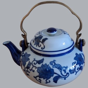 Puede incluir: Una tetera de porcelana blanca con un diseño floral azul y blanco. La tetera tiene un asa de latón y una tapa con un pomo azul.