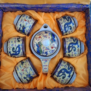 Puede incluir: Un juego de seis tazas de té y una tetera con un diseño de dragón. Las tazas y la tetera están decoradas con detalles dorados y un patrón floral azul y blanco. El juego se presenta en una caja azul y dorada.