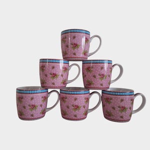 Könnte beinhalten: Sechs rosa Keramikbecher gestapelt. Jeder Becher hat ein florales Rosenmuster mit weißen Tupfen und einen blau-weiß karierten Rand. Die Becher haben graue Griffe und sind pyramidenförmig angeordnet.