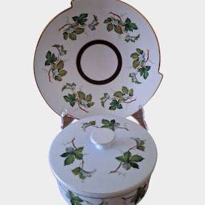 May include: A white ceramic serving dish with a lid. The dish and lid are decorated with a green and gold floral pattern. The dish has a scalloped edge and a black rim.