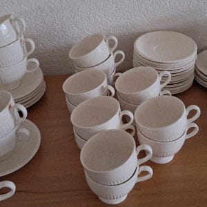 Può includere: Una collezione di tazze da tè e piattini bianchi disposti su una superficie di legno. Le tazze hanno un design a coste e sono impilate. Sono visibili anche diverse pile di piattini, che suggeriscono un servizio da tè completo.