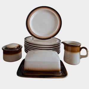 Può includere: Un set completo di stoviglie in gres composto da piatti, zuccheriera con coperchio, burriera e lattiera. Il set ha un interno bianco con bordo e dettagli marroni. I piatti sono impilati, su sfondo bianco.