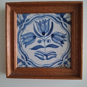 Può includere: Piastrella quadrata in ceramica incorniciata con un disegno floreale blu su sfondo bianco. La cornice in legno è marrone caldo. La piastrella presenta un fiore centrale con tre petali e altri elementi floreali.