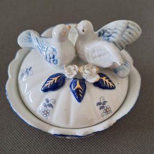 Può includere: Un piatto portaoggetti in porcellana bianca con un motivo floreale blu e oro. Due colombe in porcellana bianca con dettagli blu e dorati sono appollaiate sul bordo del piatto.