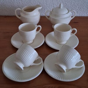 Op de afbeelding: Een crèmekleurige theeset met een melkkan, suikerpot met deksel en zes kopjes met schotels. De kopjes en schotels hebben een geribbeld ontwerp, terwijl de schotels een spiraalpatroon hebben. De set is op een houten oppervlak geplaatst.