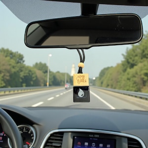 Könnte beinhalten: Ein Lufterfrischer für das Auto, der am Rückspiegel hängt. Der Lufterfrischer hat eine Holzoberseite mit dem Schriftzug "Loved Oils" und eine schwarze Unterseite mit einem weißen Tropfendesign. Das Auto fährt auf einer Autobahn.