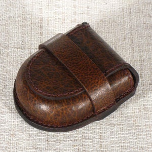 Pode incluir: Uma bolsa de moedas de couro marrom com fecho de aba e borda costurada. A bolsa está fechada e está sobre uma superfície de tecido texturizada.