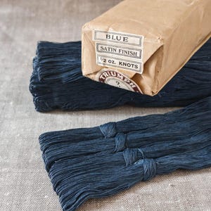 May include: A bundle of dark blue thread with a satin finish, wrapped in brown paper packaging. The label reads BLUE, SATIN FINISH, 1/2 oz. KNOTS, and TRIUMPH. The thread is organised into long, thin strands secured with small knots at regular intervals.