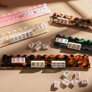 Può includere: Un set di cinque supporti rettangolari per tessere del mahjong con bordi smerlati in motivi rosa, crema, tartarugato e verde scuro. Diverse tessere bianche del mahjong con caratteri e punti rossi, verdi e neri sono disposte sui supporti.