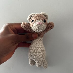 May include: A handmade crochet ferret toy in shades of beige and brown. The ferret has a light pink nose, black eyes, and outstretched arms. The toy is made with a textured yarn, giving it a soft, plush appearance. The ferret is a cute and cuddly stuffed animal.