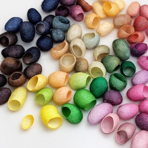 Può includere: Una collezione di bozzoli di seta ovali e colorati in varie tonalità di blu, marrone, giallo, verde, rosa e arancione. I bozzoli sono disposti su una superficie bianca, mostrando le loro trame e tonalità naturali.