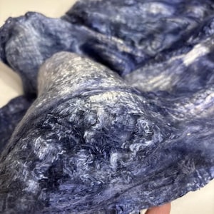 May include: A textured textile material featuring a blend of deep navy blue and shimmering silver fibers. The fabric displays a loose, fibrous weave with a soft, voluminous appearance, characterized by swirling patterns of dark and light tones.