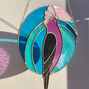 May include: A stained glass suncatcher featuring a pink and black bird with a blue and green background. The suncatcher is hanging from a silver chain.