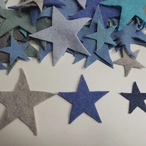 May include: An assortment of felt stars in shades of blue, grey, and teal. The stars are of different sizes, some scattered and others grouped together. The fabric has a textured appearance, and the stars are cut in a five-point design.