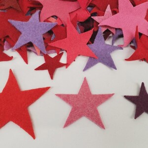 May include: Felt stars in shades of red, pink, and purple scattered on a white surface. Some stars are piled together, others are spread out. The stars vary in size, creating a visually appealing arrangement.