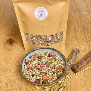 May include: A brown paper bag with a white label that says "Hammy's Tuck Shop" and a cartoon hamster holding a carrot. The bag contains a mix of dried fruits, seeds, and grains. A bowl of the same mix is on a wooden table with a few oat stalks and two brown sticks.
