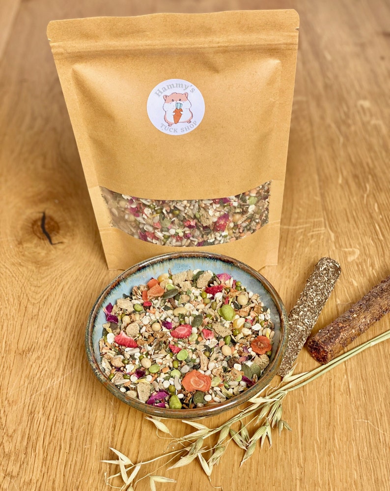 May include: A brown paper bag with a white label that says "Hammy's Tuck Shop" and a cartoon hamster holding a carrot. The bag contains a mix of dried fruits, seeds, and grains. A bowl of the same mix is on a wooden table with a few oat stalks and two brown sticks.